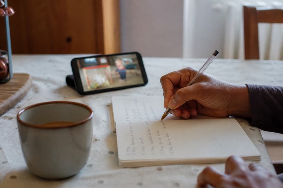 A parent writing dated notes at a kitchen table with a phone nearby, ready to take to a GP appointment.