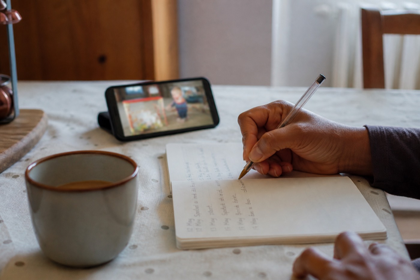 A parent writing dated notes at a kitchen table with a phone nearby, ready to take to a GP appointment.
